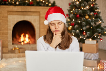 Fototapeta na wymiar Woman in Santa hat and white sweater with laptop, has thoughtful facial expression, keeping hand on chin, looks at notebook's screen doing her freelance work during Christmas holiday.