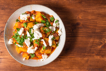 Greek vegetable stew of potatoes and vegetables in tomato sauce with chunks of soft feta cheese in a plate on a wooden table, top view, copy space