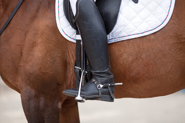 Croup of a red horse with a white saddle and a rider’s foot in a boot with a spur inserted in a...