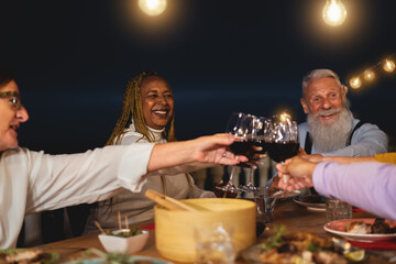 Happy multiracial senior friends toasting with red wine glasses together on house patio dinner - Elderly lifestyle people and food concept