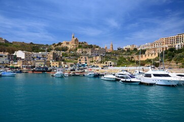 Panorama of the harbor on the island of Gozo, Malta. Boats in the harbor, church on the hill, sunny sky, Mediterranean atmosphere.