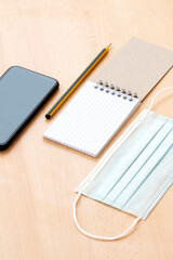 Flat lay with copy space, a face mask, a blank spiral notepad with a pen and a smart phone are arranged on a desk. To-do list concept during the Covid-19 outbreak.