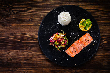 Oven baked salmon steak with white basmati rice,  lemon and fresh vegetable salad served on black stone plate on wooden table
