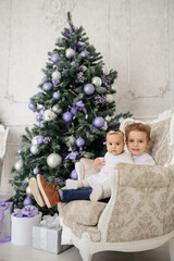 brother and sister are sitting on a vintage armchair in a white room beautiful decorated for christmas holiday with a Christmas tree and christmas presents.