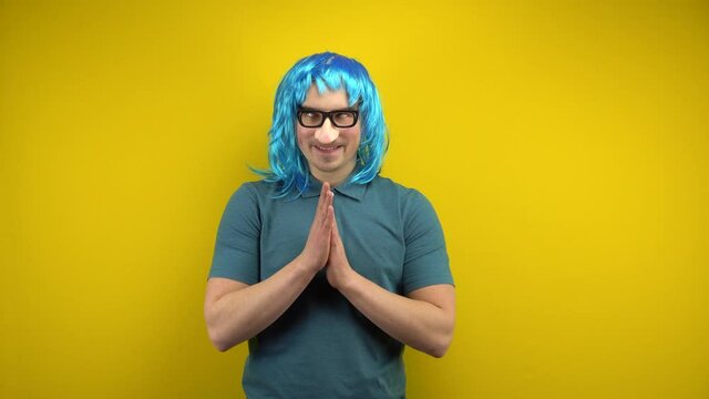 A funny young man in a female blue wig and glasses with a nose rubs his hands and looks around. Shooting in the studio on a yellow background.