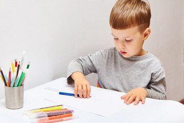 Cute little boy with blond hair draws colored pencils at home. Draws at the white table.