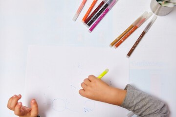 Cute little boy with blond hair draws colored pencils at home. Draws at the white table. Close up of child's hands drawing at white paper