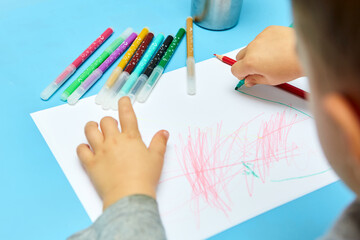 Cute little boy with blond hair draws colored pencils at home. Draws at the blue table. Close up of child's hands drawing at white paper