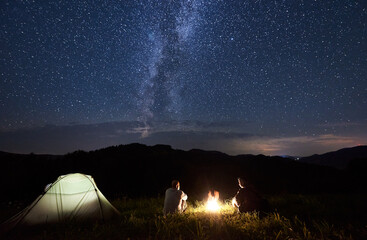 Couple of friends sitting around a campfire in the mountains in summer, enjoying incredible weather under beautiful milky way constellation. Powerful mountains under magnificent sky with bright stars