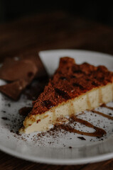 Tasty slice of coffee cheesecake covered with cacao on white plate with chocolate and caramel on dark wooden background close up