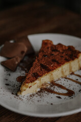 Tasty slice of coffee cheesecake covered with cacao on white plate with chocolate and caramel on dark wooden background close up