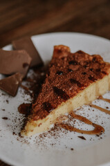 Tasty slice of coffee cheesecake covered with cacao on white plate with chocolate and caramel on dark wooden background close up