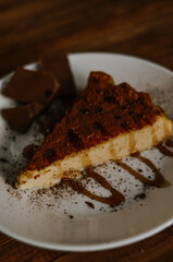 Tasty slice of coffee cheesecake covered with cacao on white plate with chocolate and caramel on dark wooden background close up