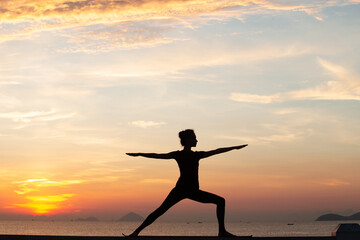 Fototapeta premium Silhouette of a young woman doing yoga near the sea at sunset.