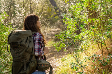 Young woman with light skin and dark hair walking through the mountain of excursion observing the trees with camping backpack, woman wears plaid shirt and jeans