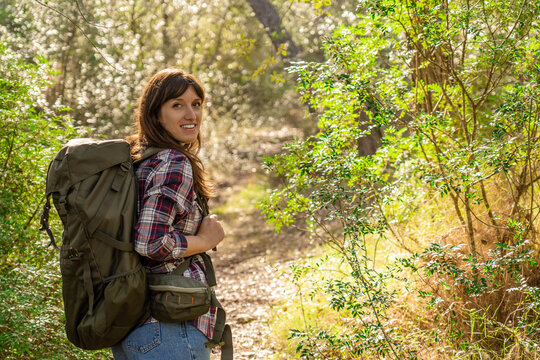 Young woman with light skin and dark hair smiling at the camera looking back outing on a mountain path in the Mediterranean landscape, Mallorca, woman wears a plaid shirt and jeans