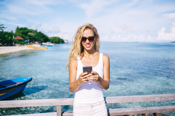 Fototapeta na wymiar Cheerful woman in sunglasses enjoying smartphone messaging with social followers communicate about Maldives vacations, smiling female tourist typing text publication for sharing to travel web blog