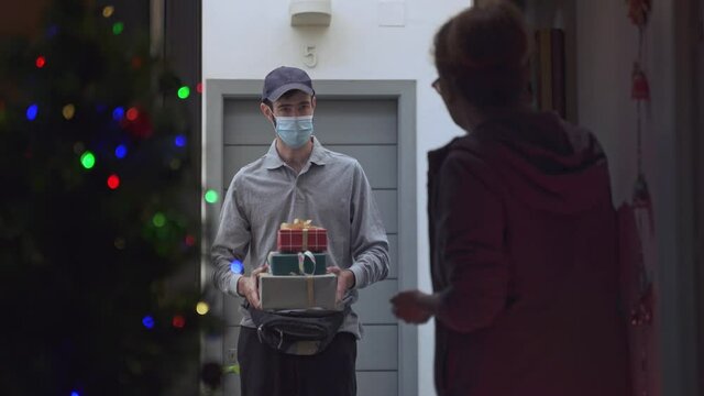 Delivery messenger giving Christmas gift boxes to a woman during coronavirus. Courier service worker bringing X-mas presents to a lady during quarantine. Social distance restrictions.