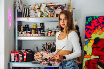 Beautiful female artist stands in a painting studio with a brush and palette in her hands and poses for the camera with a serious face. Stylish creative woman working on a picture