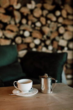 Empty coffee cup and kettle in front of velvet armchair and wood logs wall