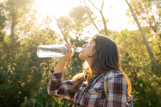 Young hiker drinking water from glass bottle with sunlight on background in forest with mediterranean landscape, woman wears plaid shirt