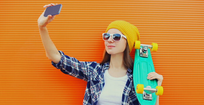 Portrait of young woman with skateboard taking selfie picture by phone over an orange background