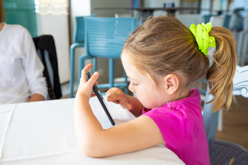 little girl with pigtail using mobile phone sitting in restaurant
