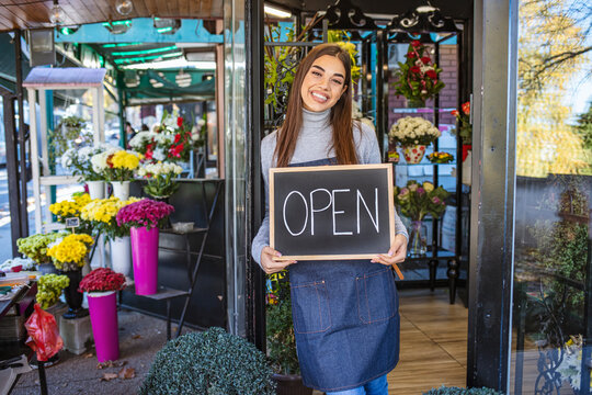 Smiling young florist in aorn holding open sign while standing in doorway of flower shop. Opening a shop. Happy florist standing in the doorway of her flower shop with an open sign.