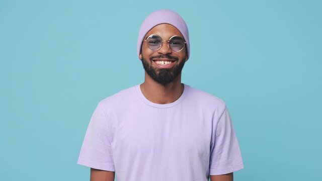 Smiling cheerful young bearded african american man 20s wearing violet t-shirt hat glasses posing isolated on blue turquoise color background in studio. People lifestyle concept. Looking at camera