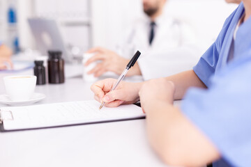 Medical woman practitoner writing notes on clipboard wearing blue uniform during conference with coworkers. Clinic expert therapist talking with colleagues about disease, medicine professional.
