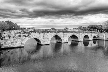 Black and white view of the Tiberius Bridge or Ponte di Augusto, a Roman bridge in Rimini, Italy, under a dramatic sky