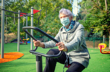 A grey-haired elderly woman in a face mask is exercising on a spinning cycle trainer in her local outdoor gym circuit.