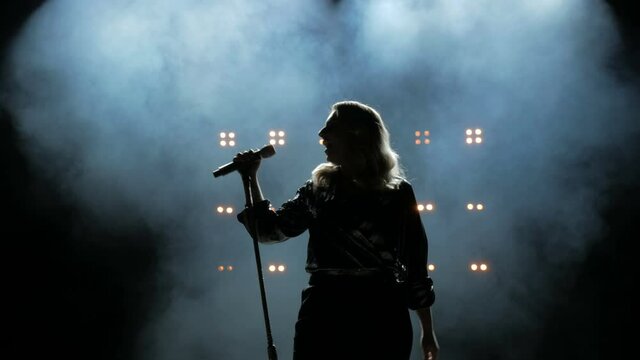 Silhouette of a girl comes to the microphone on the stage against a background of bright yellow lights