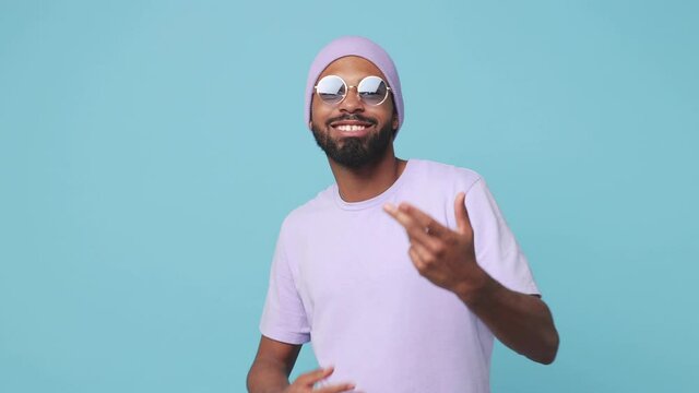 Smiling cheerful young african american man in violet t-shirt hat glasses isolated on blue background studio. People lifestyle concept. Dancing clenching fists point finger camera showing victory sign