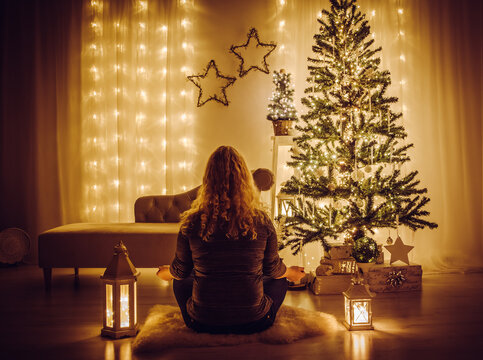 Woman sit on sheepskin rug and meditating. Tranquil relaxing Christmas Eve concept. Decorated Christmas tree with icicles and snowflake ornaments and party lights. Star shape led curtains hanging.