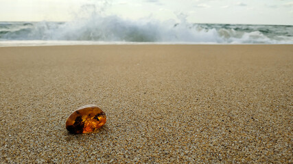 Natural piece of sparkling orange Baltic amber, glowing in the sun, lies on the smooth sand on the seashore, a wave with foam wants to cover it. 