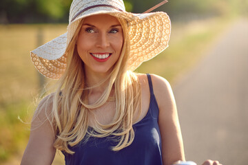 Attractive middle-aged blond woman in a wide brimmed straw sunhat and blue summer top smiling at the camera as she glances aside on a sunny rural road