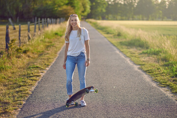 Attractive athletic blond woman with her skateboard backlit by the evening sun on a rural road between fields smiling at the camera