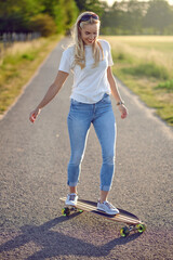 Fit active middle-aged woman playing on her skateboard approaching the camera along a narrow rural road with a happy smile backlit by the evening sun