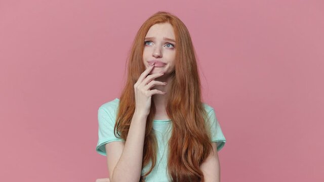 Confused puzzled ginger young woman in blue turquoise t-shirt posing isolated on pink background studio. People lifestyle concept Looking camera aside ask what spreading hands put hand prop up on chin