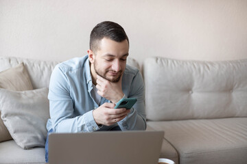 Businessman working at home using laptop and mobile phone. Home office. Young man sitting relaxed on sofa with laptop and smartphone. 