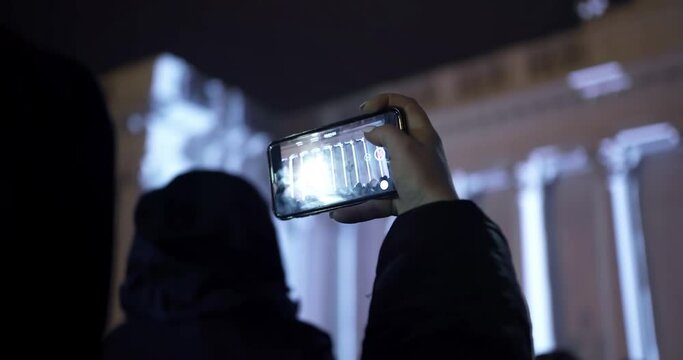 Woman in Warm Clothes Filming Night Light Show on Phone. Video Mapping Projection on the Building.