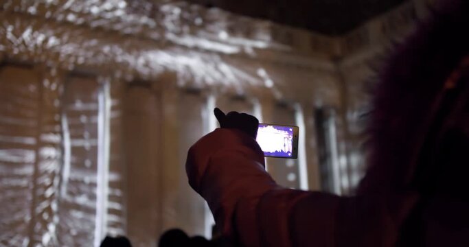 Woman in Warm Clothes Filming Night Light Show on Phone. Video Mapping Projection on the Building.