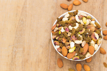 A variety of nuts are in a white bowl on the wooden background at the right of the image, top view, flay lay, top-down.