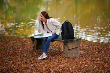 Beautiful young college student sits alone on campus in fall leaves near small pond - writing in notebook and laughing