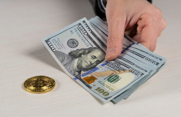 Male hand holds one hundred dollar bills near the gold bitcoin coin.