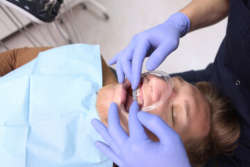 Fototapeta premium A young man installs braces at the dental clinic. Unrecognizable doctor orthodontist. Hands in protective gloves. Health care concept.