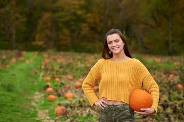 Adorable young woman in yellow crop sweater and camouflage pants stands in a pumpkin patch on farm - autumn holidays thanksgiving or Halloween themed - holding pumpkin against hip