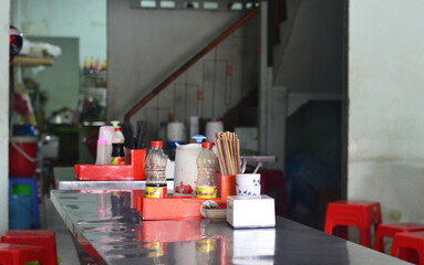 A metal table in a Vietnamese canteen, sauces and chopsticks are on the table, red stools stand...
