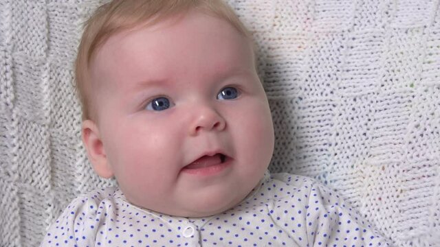 A cute little blue-eyed baby on a white knitted blanket is laughing at the camera